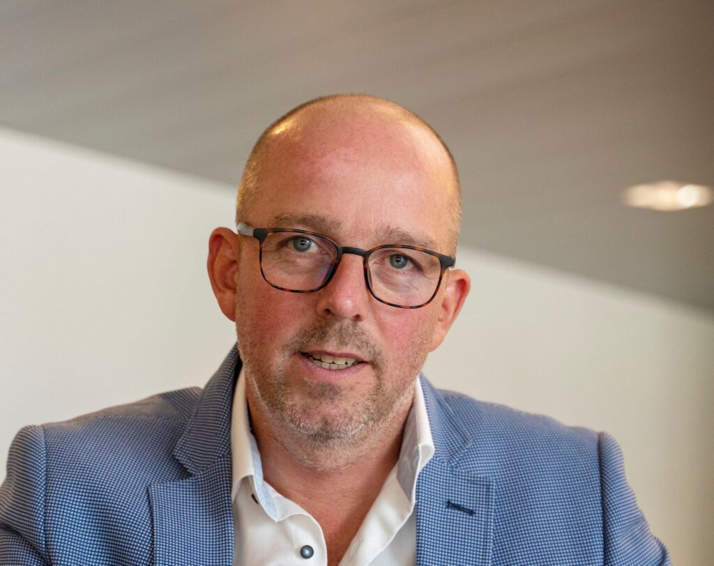 Een docent in pak met bril poseert voor de camera tijdens een les praktijkmanagement in de huisartsenzorg bij Sitis.